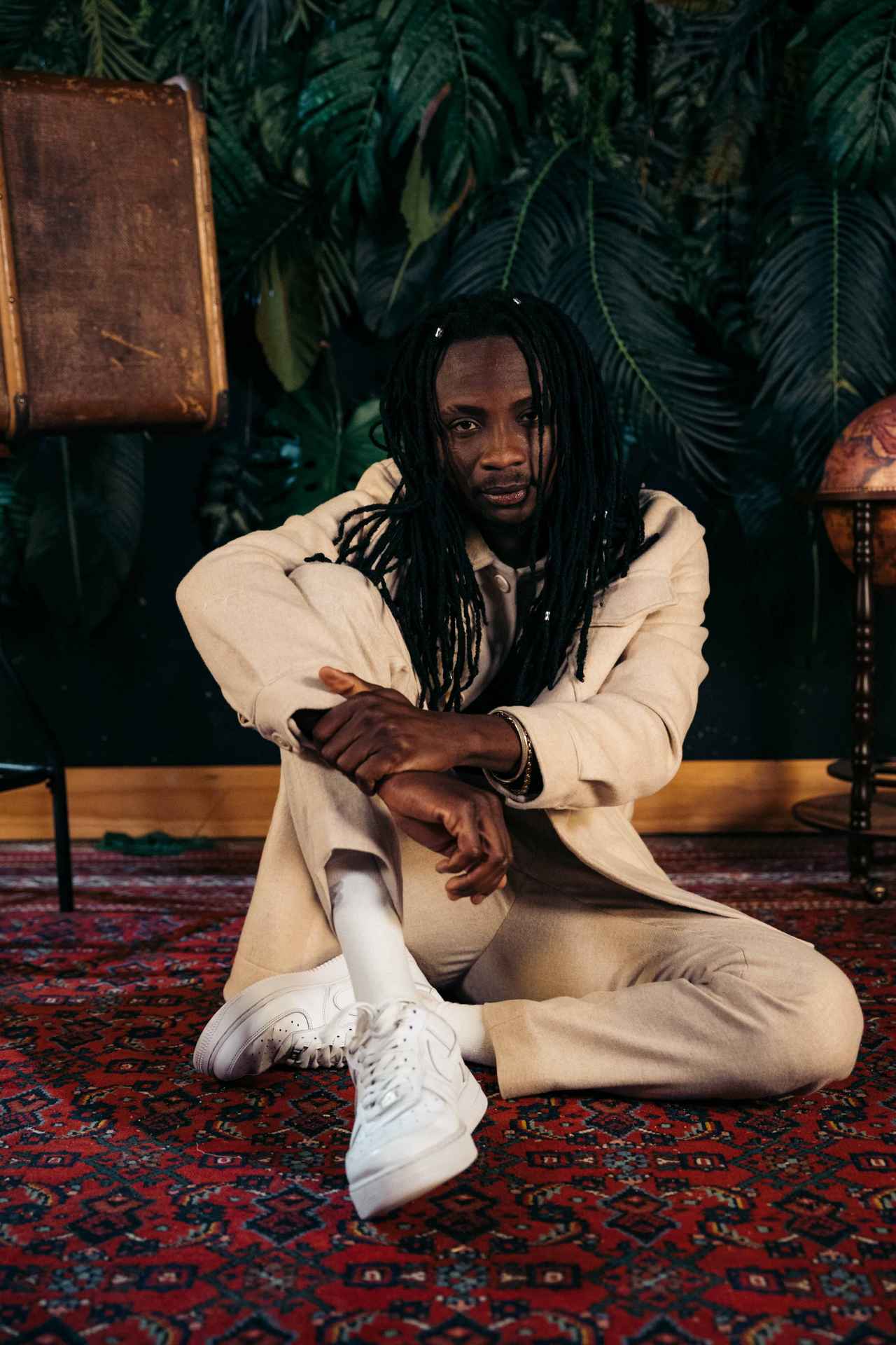 Afrobeat Artist Fofana Jo sitting on the ground in front of a dark wall and plants contrasting his bright clothes